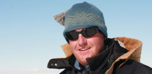 A person wearing a blue knit hat with a tassel, dark sunglasses, and a thick black winter coat stands outdoors under the clear blue sky—ready for an Antarctica adventure with Antarctic Logistics and Expeditions.