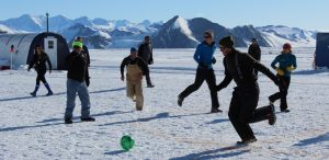 A group of people play soccer on snow in Antarctica, with snowy peaks and tents in the background. Some chase a green ball while others watch, creating a bright and lively scene supported by Antarctic Logistics and Expeditions.