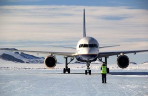 A commercial airplane is parked on a snowy runway at the South Pole in Antarctica, with mountains beyond. A person in a reflective vest, part of Antarctic Logistics and Expeditions, stands facing the aircraft, guiding or inspecting it.