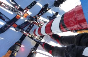A close-up view of several skiers at a starting gate in Antarctica, skis lined up side by side on the snow, ready to race. A red and white striped gate pole stands in the foreground, hinting at Antarctic Logistics and Expeditions near the South Pole.