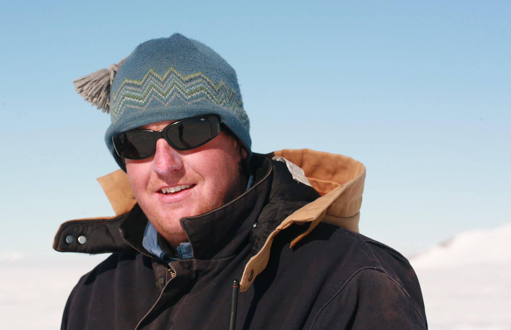 A person wearing a blue knit hat with a pom-pom, dark sunglasses, and a thick brown winter coat stands outside in a snowy, sunlit landscape—ready for Antarctica with Antarctic Logistics and Expeditions gear.