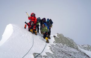 A group of climbers wearing cold weather gear and helmets stand together on a snowy mountain ridge in Antarctica, roped together and posing for a photo with ice axes against a gray sky.