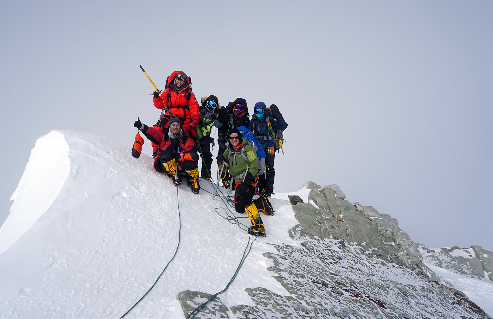 A group of climbers wearing cold weather gear and helmets stand together on a snowy mountain ridge in Antarctica, roped together and posing for a photo with ice axes against a gray sky.
