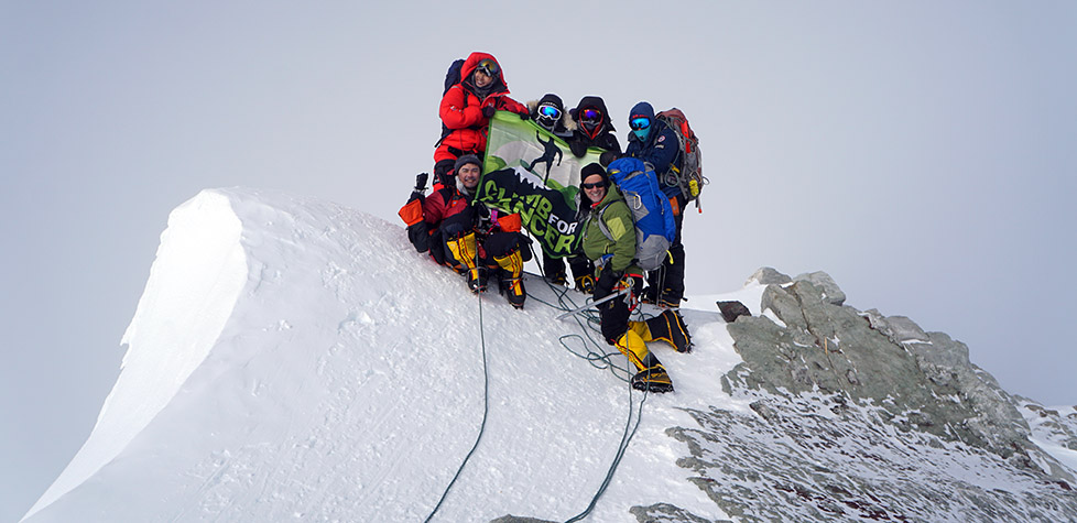 A group of climbers in winter gear stand on a snowy mountain summit in Antarctica, smiling and holding a green and black flag. Roped together, they showcase the spirit of Antarctic Logistics and Expeditions under a gray sky.