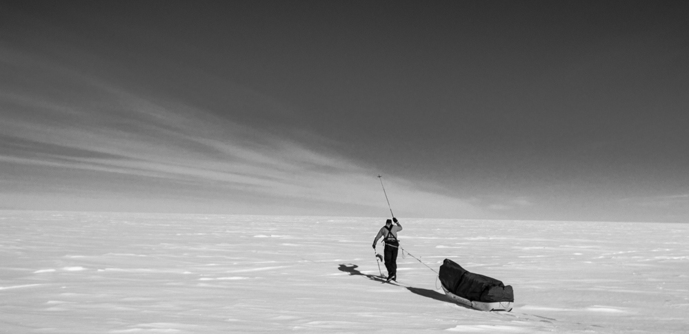 A lone person in winter gear pulls a sled across a vast, snowy Antarctic landscape under a clear sky, with long shadows stretching out behind them—showcasing the spirit of Antarctic Logistics and Expeditions.