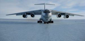 A large, four-engine cargo aircraft operated by Antarctic Logistics and Expeditions is parked on an icy, snow-covered runway in Antarctica under a partly cloudy sky.