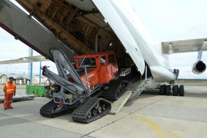 A tracked utility vehicle is being unloaded from the cargo hold of a large airplane via a ramp in Antarctica. A worker in a high-visibility jacket and helmet, part of Antarctic Logistics and Expeditions, stands nearby on the tarmac.