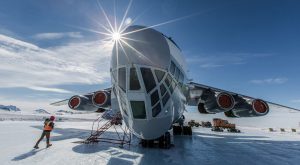 A large aircraft is parked on a snowy South Pole runway under a clear blue sky, with the sun shining brightly above its nose. A person in winter clothing walks nearby and vehicles from Antarctic Logistics and Expeditions sit in the background.