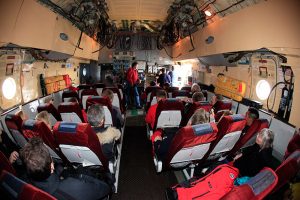 Interior of an Antarctic Logistics and Expeditions aircraft with rows of red and gray seats occupied by passengers, some looking forward, others talking. Exposed wiring and equipment line the ceiling; oval windows reveal views en route to Antarctica.