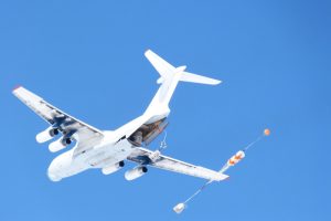 A large white cargo aircraft flies against a clear blue sky over Antarctica with its rear cargo door open, deploying a parachute and container load in mid-air for Antarctic Logistics and Expeditions.