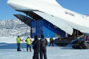 Several people in winter clothing unload a large blue shipping container from the open cargo hold of a white transport plane on the snowy, icy landscape of Antarctica, with mountains in the background—a scene typical for Antarctic Logistics and Expeditions.