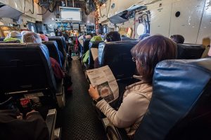 Passengers sit in rows inside an aircraft cabin, with one person in the foreground reading a newspaper. The view is toward the cockpit, and various equipment and bags—typical of Antarctic Logistics and Expeditions flights—are visible overhead.