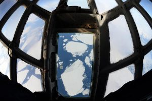 View from inside an aircraft cockpit looking down through a window at floating icebergs in the ocean below near Antarctica, with multiple glass panels and metal frames visible—an experience made possible by Antarctic Logistics and Expeditions.