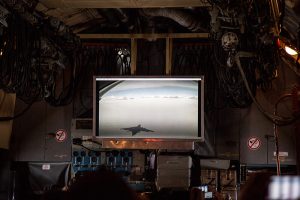 A screen inside an aircraft displays the shadow of a jet flying over snow-covered terrain near Antarctica's mountains; aircraft equipment and cables surround the screen, highlighting Antarctic Logistics and Expeditions in action.