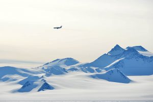 A solitary airplane flies above snow-covered mountains and the vast icy landscape of Antarctica under a pale sky, evoking the spirit of Antarctic Logistics and Expeditions.