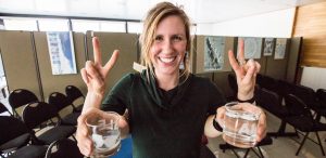 A smiling person holds two glasses of ice water, each hand making a peace sign. They stand in a room with black chairs and Antarctic Logistics and Expeditions posters on the walls in the background.
