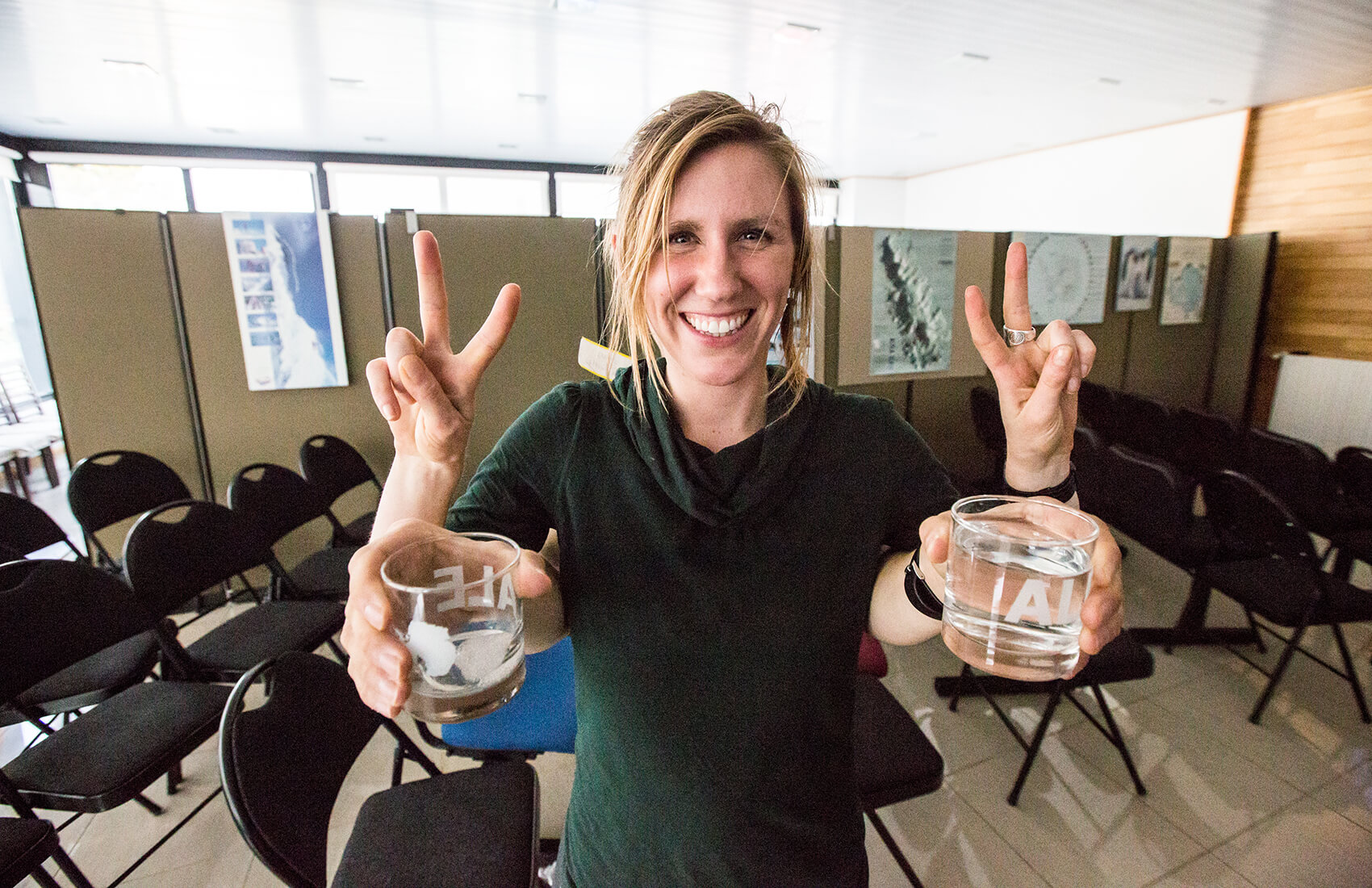 A smiling person stands indoors holding two clear glasses of water labeled H2O, making a peace sign with both hands. Chairs and Antarctic Logistics and Expeditions display boards are visible in the background.