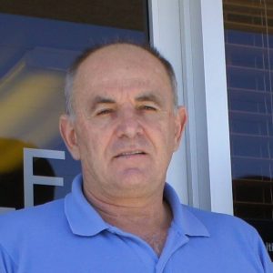 A middle-aged man with short gray hair wearing a light blue polo shirt stands outdoors in front of a building with large windows, possibly associated with Antarctic Logistics and Expeditions in Antarctica.