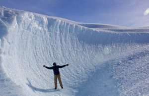 A person wearing a jacket and pants stands with arms outstretched on a curved, icy surface surrounded by towering, textured blue ice under a clear sky in Antarctica, showcasing the adventure of Antarctic Logistics and Expeditions.