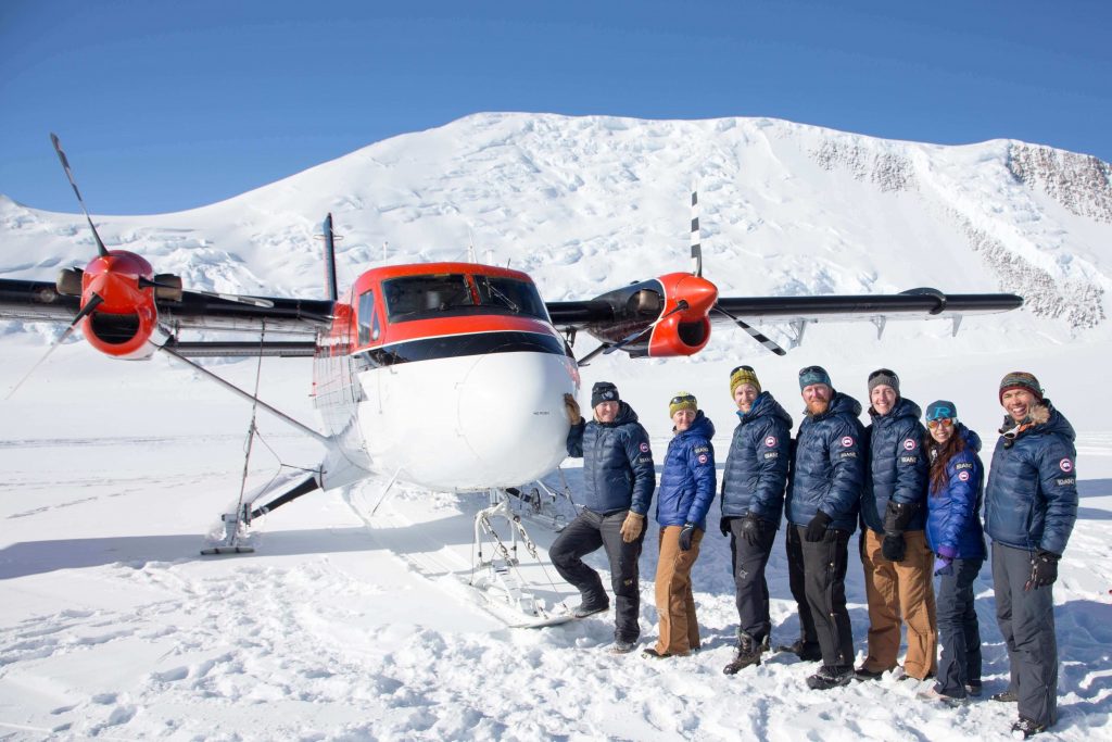 Seven people in winter jackets stand in a row, smiling, in front of a small plane on snowy ground in Antarctica, with majestic mountains behind them—a classic scene from an Antarctic Logistics and Expeditions adventure under clear blue skies.