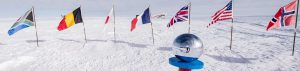 A reflective silver sphere on a blue-and-red stand sits in the snow at the South Pole, surrounded by national flags including the United States, United Kingdom, Norway, and others, with an Antarctic landscape and an airplane from Antarctic Logistics and Expeditions nearby.