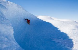 A person dressed in bright winter clothing climbs up a steep, snowy slope under a clear blue sky in Antarctica, surrounded by smooth, white snow-covered terrain.