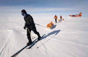 Three people in winter gear pull sleds across the snowy, flat landscape of Antarctica on skis. In the background, an Antarctic Logistics and Expeditions airplane is parked beneath a clear sky.