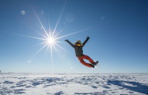 A person in a winter coat and orange pants jumps joyfully on a snowy Antarctic landscape under a bright sun, capturing the spirit of adventure enabled by Antarctic Logistics and Expeditions.