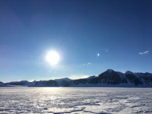 The sun shines brightly over a snowy, frozen landscape near the South Pole, with distant dark, rugged mountains under a clear blue sky.