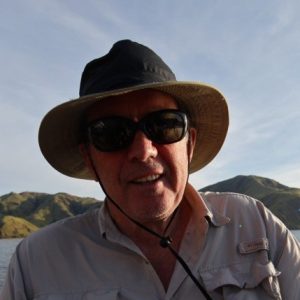 A man wearing a wide-brimmed hat and sunglasses smiles outdoors with hills and a blue sky in the background. He is dressed in a light-colored, button-up shirt, ready for an Antarctic Logistics and Expeditions adventure.