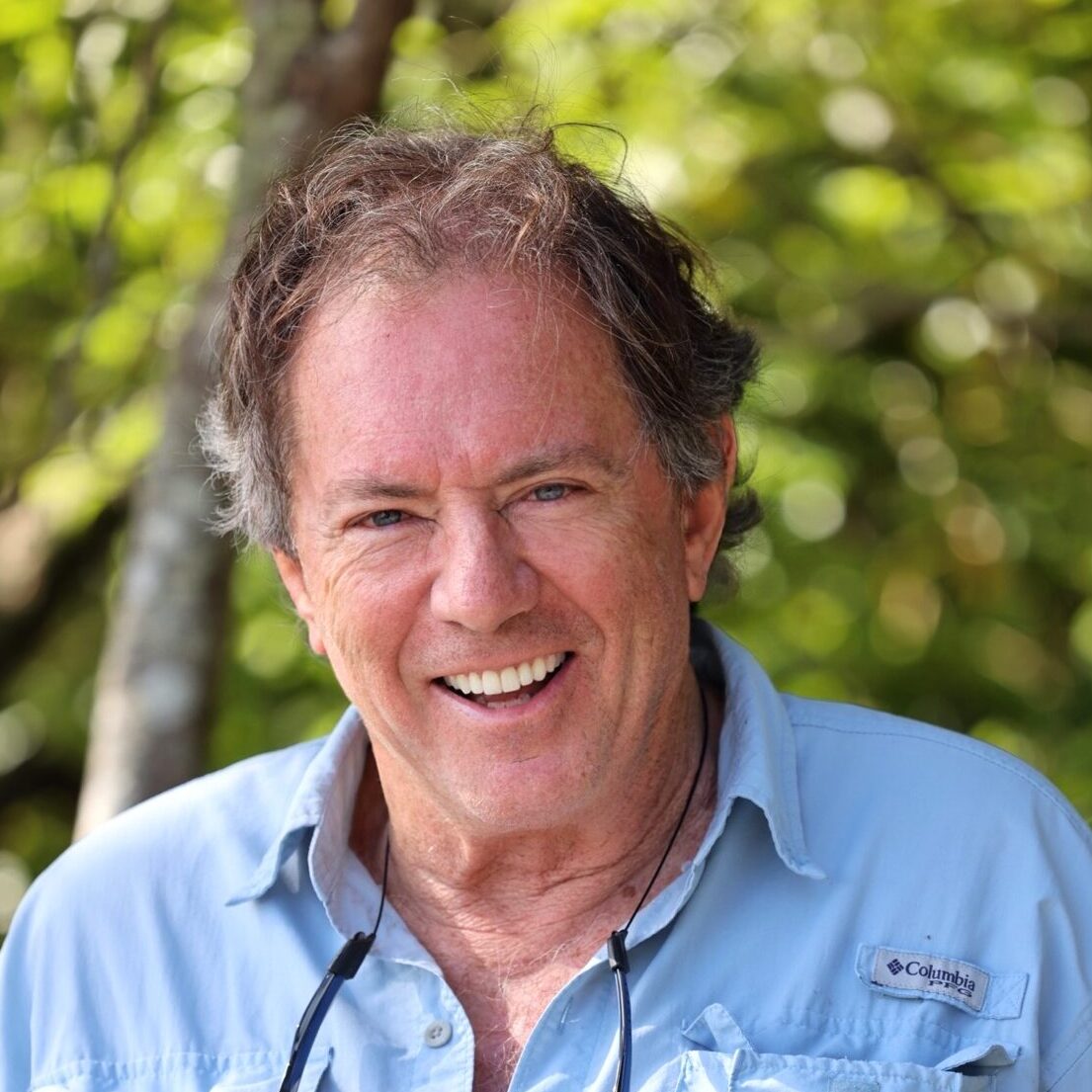 A smiling older man with graying hair wears a light blue Columbia shirt outdoors, trees and greenery blurred behind him. His experience with Antarctic Logistics and Expeditions fuels his stories of adventures near the South Pole.