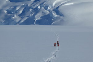 Four people in bright clothing walk in single file, leaving a trail in the snow toward rugged, snow-covered mountains under a clear Antarctic sky.