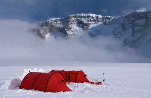Two bright red tents from Antarctic Logistics and Expeditions sit on a snowy landscape in Antarctica, with snow shovels nearby and a backdrop of snow-covered mountains and drifting clouds.