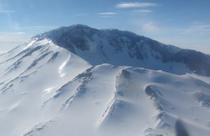Snow-covered volcanic mountain peak with sharp ridges and deep shadows under a clear blue sky in Antarctica. Sunlight highlights the smooth, white surface and rugged features—perfect for Antarctic Logistics and Expeditions enthusiasts.