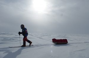A person in cold weather gear skis across a snowy, flat landscape in Antarctica, pulling a red sled behind them under a cloudy sky with the sun barely visible—a classic scene of Antarctic Logistics and Expeditions.