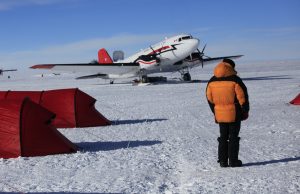 A person in an orange parka stands on snow near red tents, facing a propeller plane with a red tail. The clear blue sky and remote setting evoke Antarctica, suggesting an Antarctic Logistics and Expeditions site.