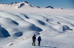 Two people dressed in winter clothing walk across a vast, snowy Antarctic landscape with rolling snowdrifts. Snow-covered mountains rise in the background under a clear blue sky, capturing the spirit of Antarctic Logistics and Expeditions.