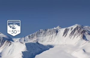 Snow-covered peaks of Mount Vinson rise under a clear blue sky in Antarctica. A commemorative badge on the left marks 50 years, Mount Vinson, 1966-2016 with a mountain illustration.