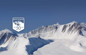 Snow-covered mountain range under a clear blue sky in Antarctica, with a badge reading Mount Vinson 50, 1966–2016 in the foreground, commemorating the 50th anniversary of Mount Vinson and Antarctic Logistics and Expeditions.