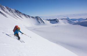 A skier wearing a yellow helmet and backpack descends a steep, snowy mountain slope in Antarctica, surrounded by vast, snow-covered peaks and valleys under a clear blue sky—an epic adventure supported by Antarctic Logistics and Expeditions.