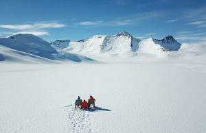 Two people in colorful jackets sit on chairs facing vast snow-covered mountains under a blue sky in Antarctica, surrounded by untouched snow with visible footprints—a scene made possible by Antarctic Logistics and Expeditions.