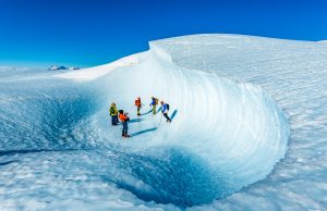 A group of people in colorful winter gear walk with ice axes and crampons on a deep, curved, icy formation in Antarctica’s bright snowy landscape under a clear blue sky, exploring terrain with Antarctic Logistics and Expeditions.