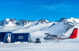 A small airplane and a blue tent structure sit on a snowy landscape in Antarctica with snowy mountains in the background under a clear blue sky. A snowmobile, used for Antarctic Logistics and Expeditions, is parked near the tent.