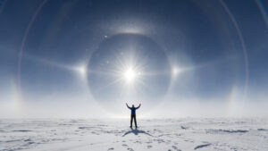 A person stands on a snowy landscape at the South Pole with arms raised, silhouetted against a bright sun surrounded by a striking halo and light rings in Antarctica’s clear, blue sky—an adventure with Antarctic Logistics and Expeditions.