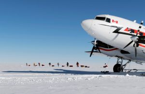 A large white airplane with a Canadian flag on the tail is parked on a snowy landscape in Antarctica. In the background, several people with Antarctic Logistics and Expeditions pull sleds across the snow under a clear blue sky.