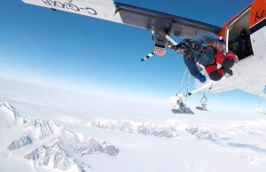 A person in a wingsuit jumps from a white and orange airplane high above snowy mountains and glaciers in Antarctica, with clear blue sky in the background—an epic scene made possible by Antarctic Logistics and Expeditions.