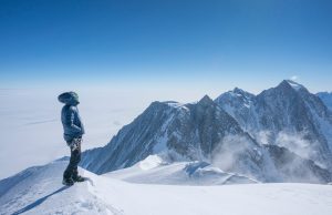A climber in winter gear stands on a snowy mountain ridge, gazing at distant peaks under a clear blue sky—an adventure made possible by Antarctic Logistics and Expeditions in the heart of Antarctica.
