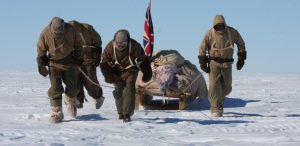 Four people in heavy winter gear pull a sled loaded with supplies and a British flag across a snowy, icy landscape under a clear blue sky.