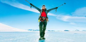 A person dressed in winter gear stands on ice with arms raised, holding trekking poles, smiling under a bright blue sky with snow-covered mountains in the background.