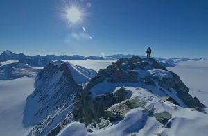 A person stands on a snow-covered mountain peak under a bright sun, with rugged mountains and vast icy plains stretching into the distance beneath a clear blue sky.